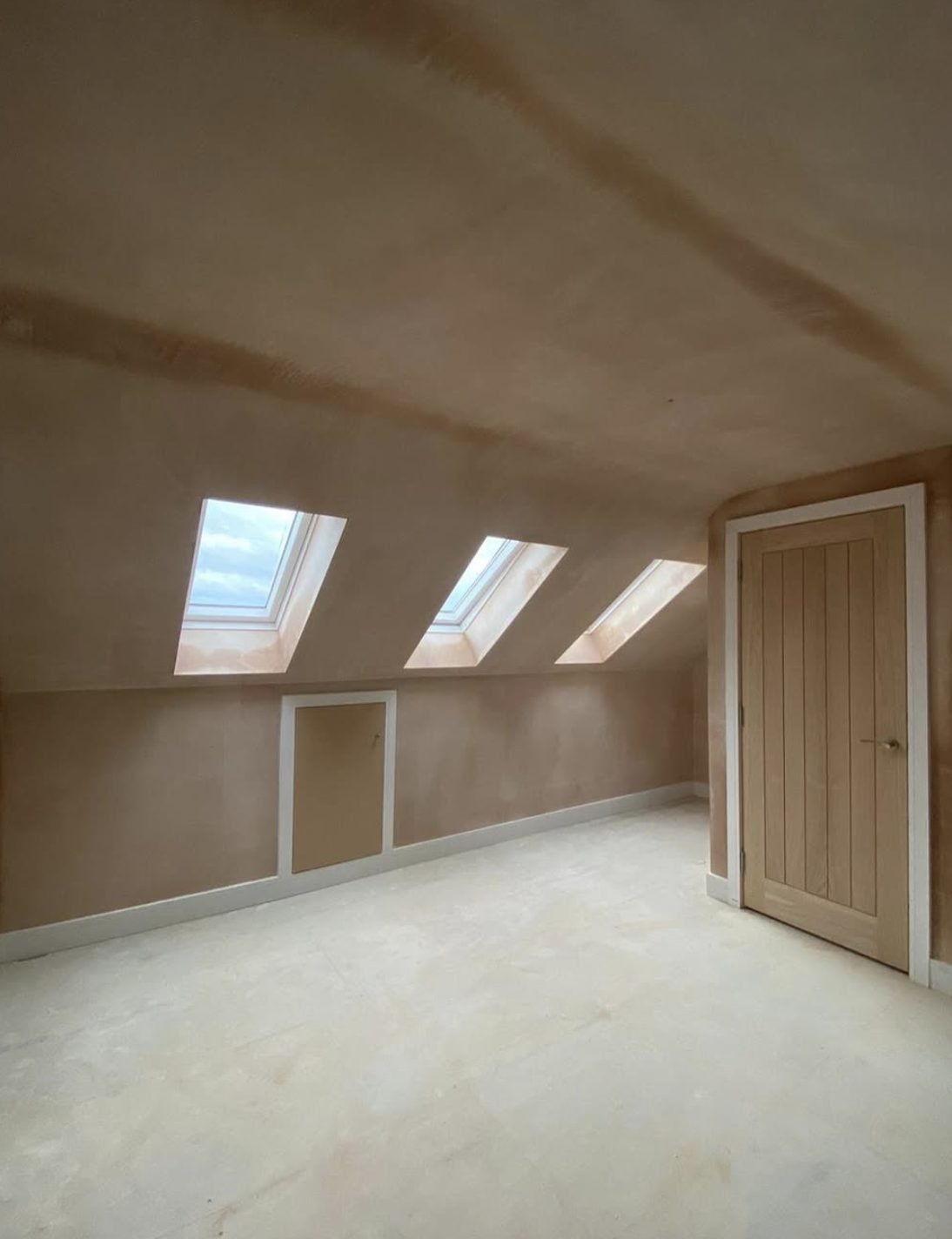 Loft conversion mid-build: freshly plastered room with three rooflights, eaves storage hatch and oak door